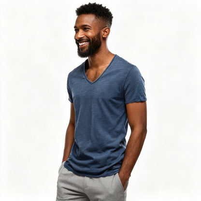 A smiling man with a beard wearing a heathered slate blue v-neck t-shirt and light gray pants, standing against a plain white studio background. - Fashlot