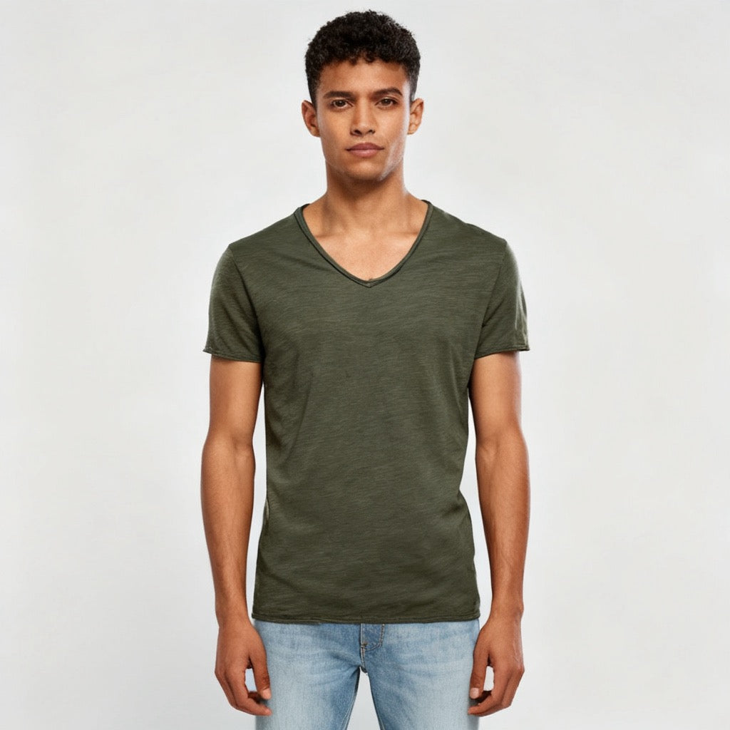 A man wearing an olive green v-neck t-shirt and light blue jeans, standing against a light gray studio background. - Fashlot