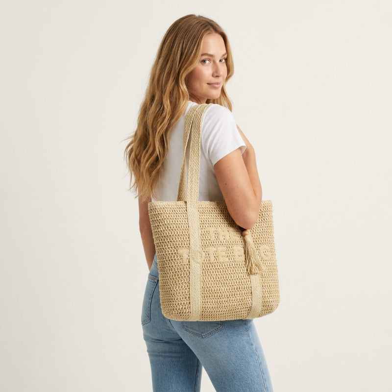 A woman with long wavy light brown hair is shown from a side-back angle, wearing a white t-shirt and blue jeans while carrying a large woven straw tote bag over her shoulder, against a plain off-white studio background. - Fashlot