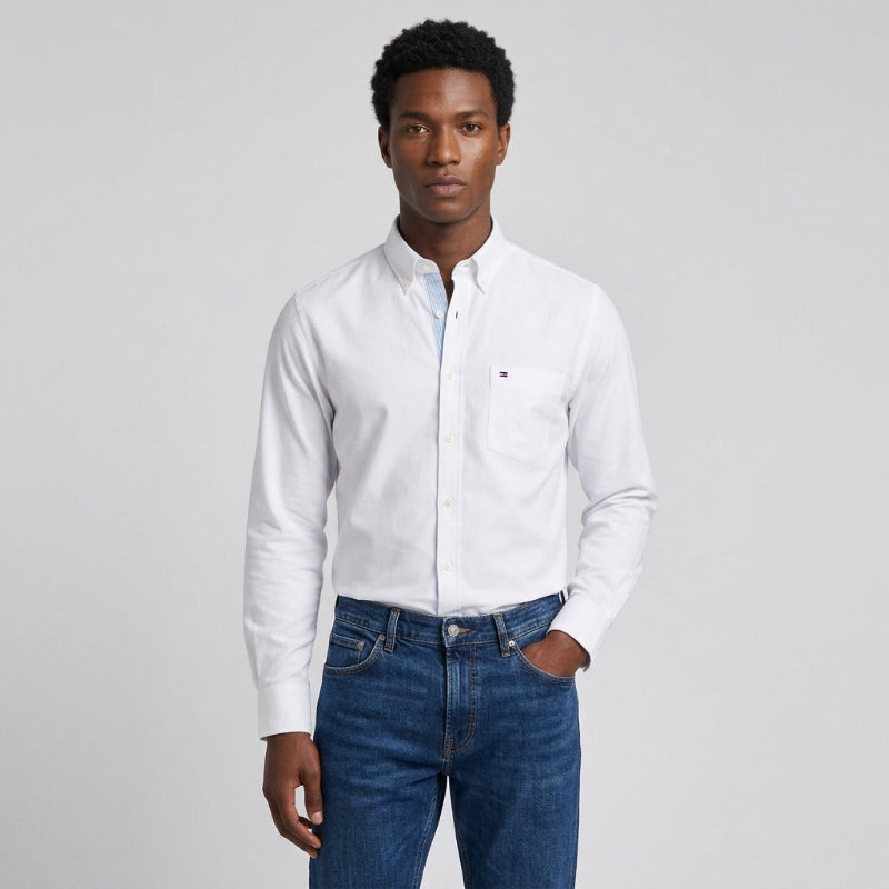 A man wearing a white long-sleeve button-down shirt with a small chest pocket and blue jeans, standing against a light gray studio background. - Fashlot