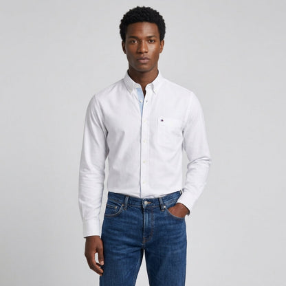 A man wearing a white long-sleeve button-down shirt with a small chest pocket and blue jeans, standing against a light gray studio background. - Fashlot