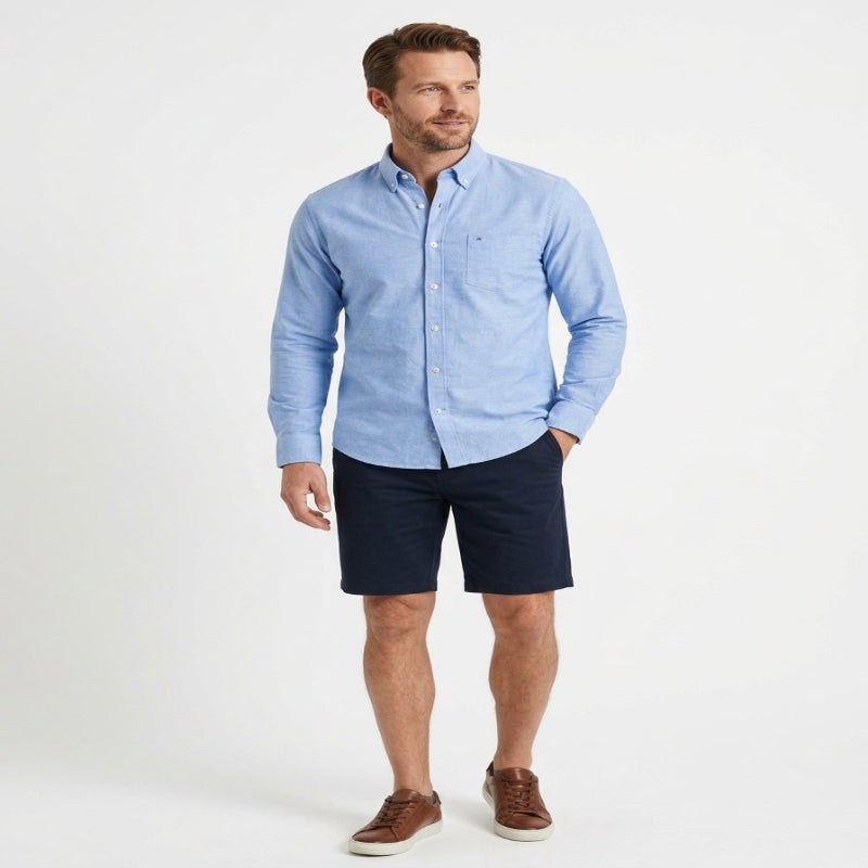 A man modeling a light blue long-sleeve button-down shirt paired with dark navy shorts and brown leather sneakers, standing against a plain white studio background. - Fashlot