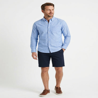A man modeling a light blue long-sleeve button-down shirt paired with dark navy shorts and brown leather sneakers, standing against a plain white studio background. - Fashlot