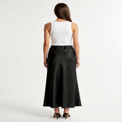 A woman shown from the back wearing a white sleeveless top and a long black satin-like skirt, standing against a plain light gray studio background. - Fashlot