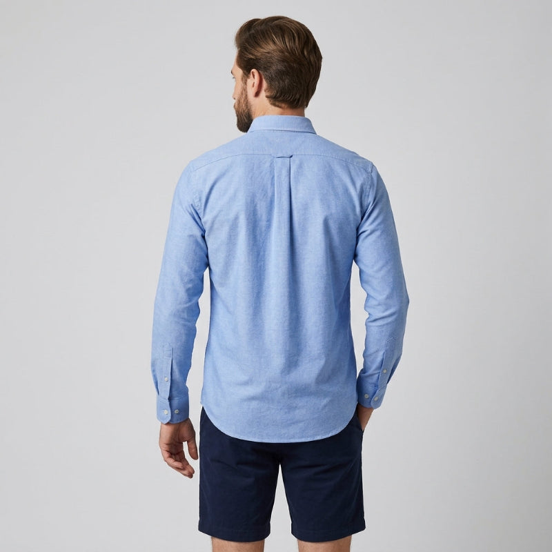 A man viewed from the back wearing a light blue long-sleeve button-down shirt and dark navy shorts, standing against a neutral light gray background. - Fashlot 