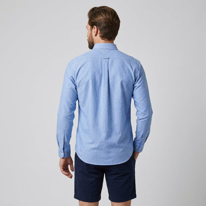 A man viewed from the back wearing a light blue long-sleeve button-down shirt and dark navy shorts, standing against a neutral light gray background. - Fashlot 