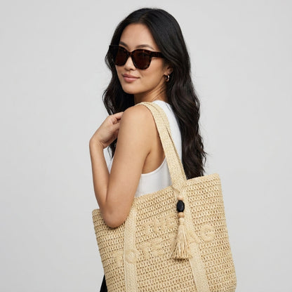 A woman wearing sunglasses and a white tank top carries a large woven straw tote bag with tassel details against a light gray studio background. - Fashlot