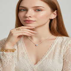 A close-up of a woman with long light brown hair wearing a delicate silver necklace with a clear crystal pendant, a gold ring, and stacked gold bracelets, paired with a cream lace top against a light gray background. - Fashlot