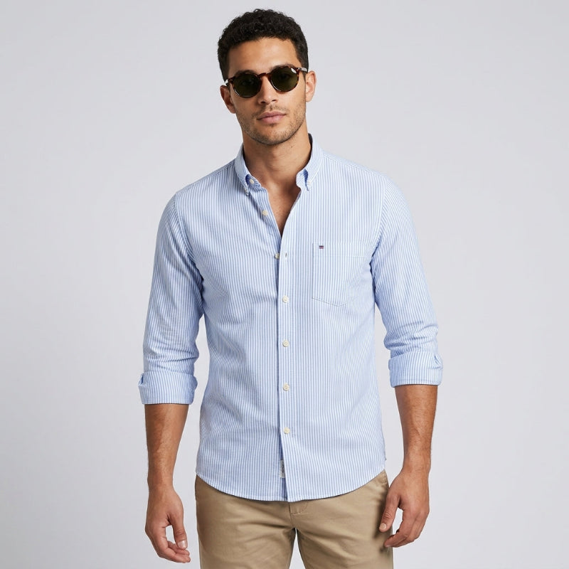 A man wearing a light blue and white vertically striped long-sleeve button-down shirt, paired with khaki pants and sunglasses, standing against a light gray studio background. - Fashlot 