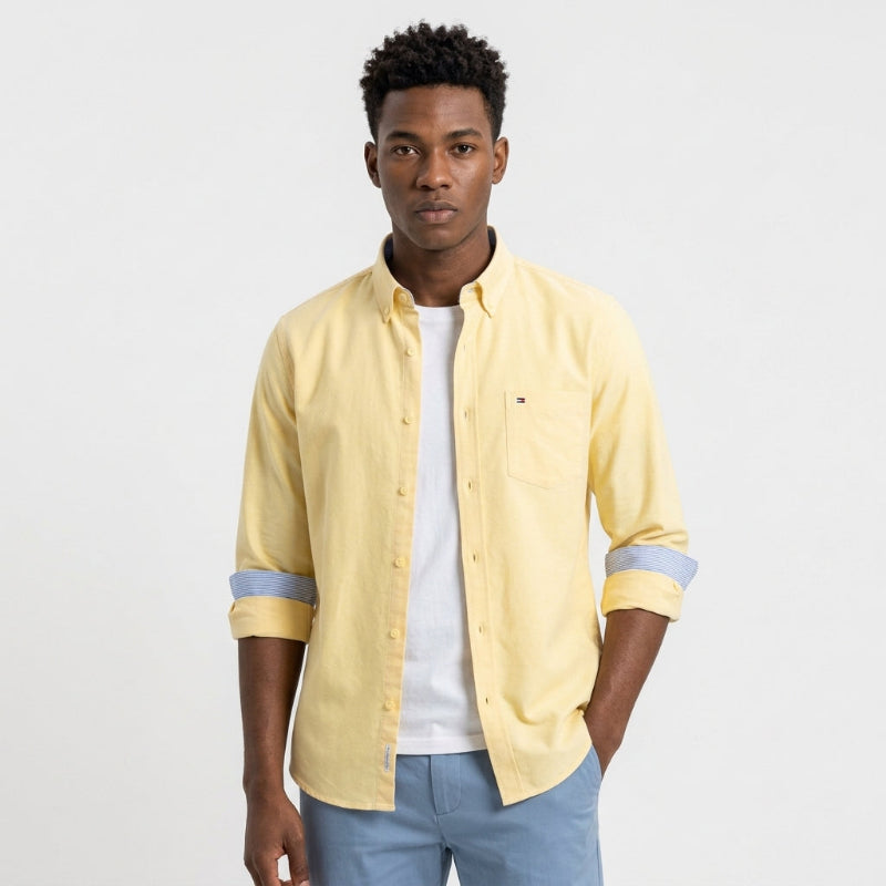 A man wearing a pale yellow long-sleeve button-down shirt with light blue rolled cuffs, layered over a white t-shirt and paired with light blue jeans, against a plain white studio background. Fashlot