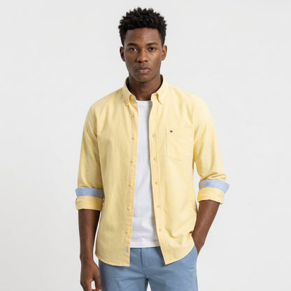 A man wearing a pale yellow long-sleeve button-down shirt with light blue rolled cuffs, layered over a white t-shirt and paired with light blue jeans, against a plain white studio background. Fashlot