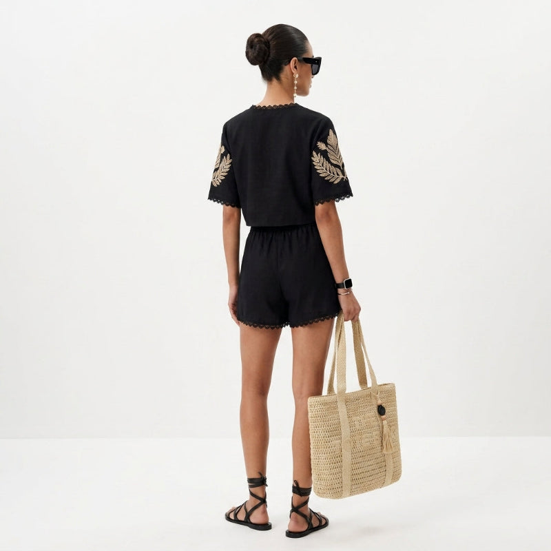 A woman shown from the back wearing a black short-sleeve romper with gold embroidered leaf details on the sleeves, holding a large beige woven tote bag against a plain white studio background. - Fashlot