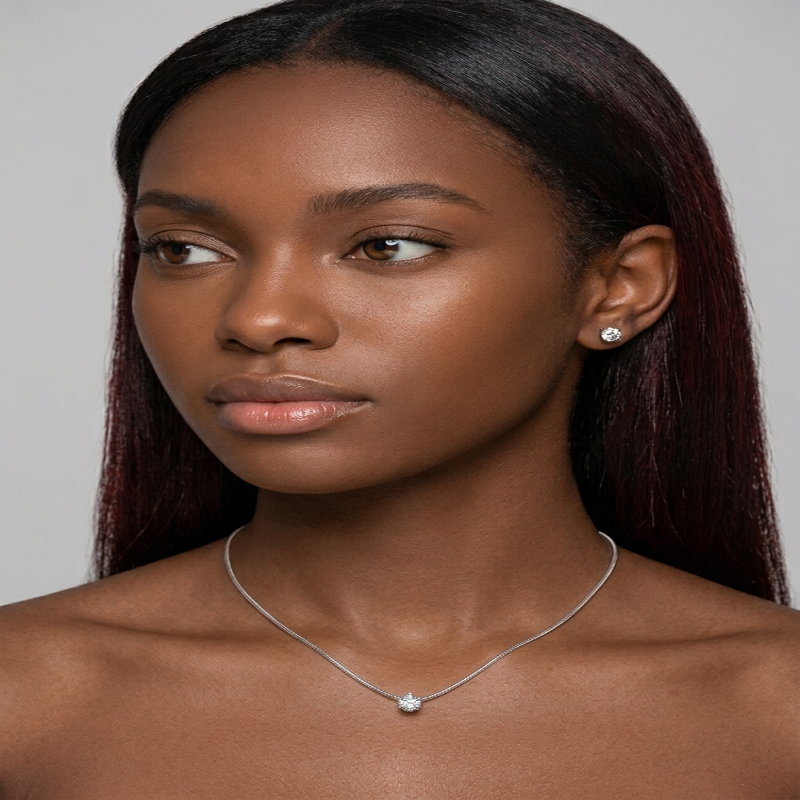 A close-up portrait of a woman with long dark hair wearing a delicate silver chain necklace with a small round clear stone pendant and a matching stud earring, against a plain light gray background. - Fashlot