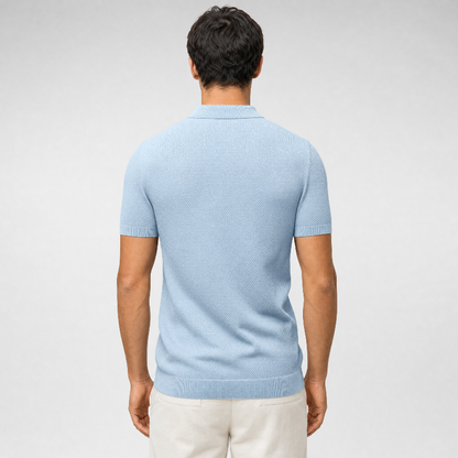 A man viewed from the back wearing a light blue short-sleeve knit polo shirt and off-white trousers, set against a neutral light gray studio background. - Fashlot