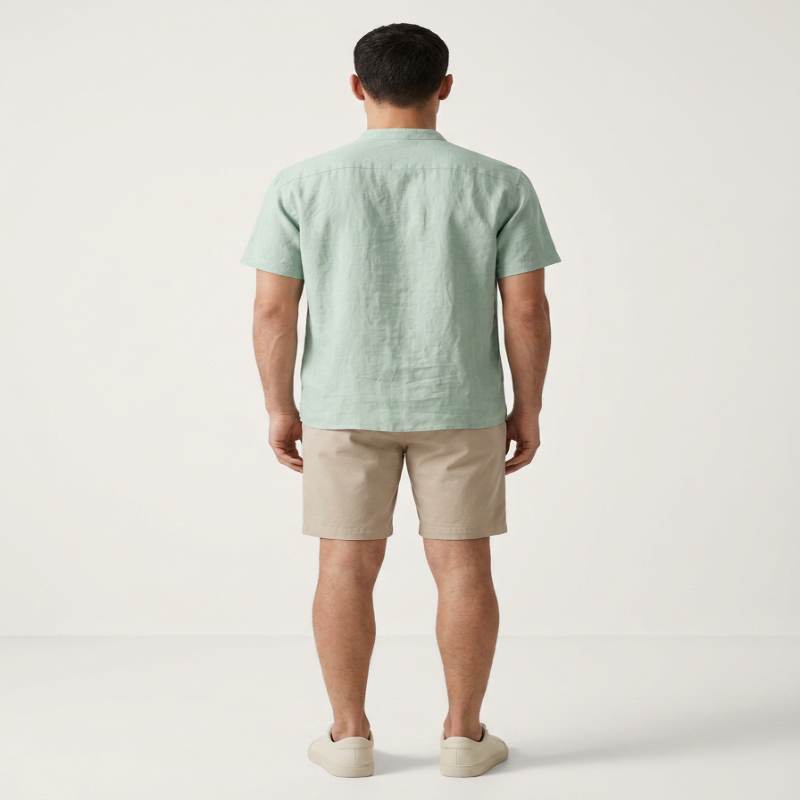 A man viewed from the back wearing a light sage green short-sleeve linen shirt, beige shorts, and off-white slip-on shoes, standing against a plain off-white studio background. - Fashlot