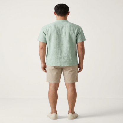 A man viewed from the back wearing a light sage green short-sleeve linen shirt, beige shorts, and off-white slip-on shoes, standing against a plain off-white studio background. - Fashlot