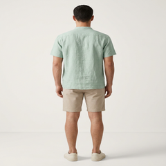 A man viewed from the back wearing a light sage green short-sleeve linen shirt, beige shorts, and off-white slip-on shoes, standing against a plain off-white studio background. - Fashlot