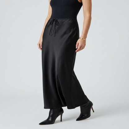 A model wearing a black satin maxi skirt with a drawstring waist, paired with a black sleeveless top and black pointed-toe heeled ankle boots, against a light gray studio background. - Fashlot